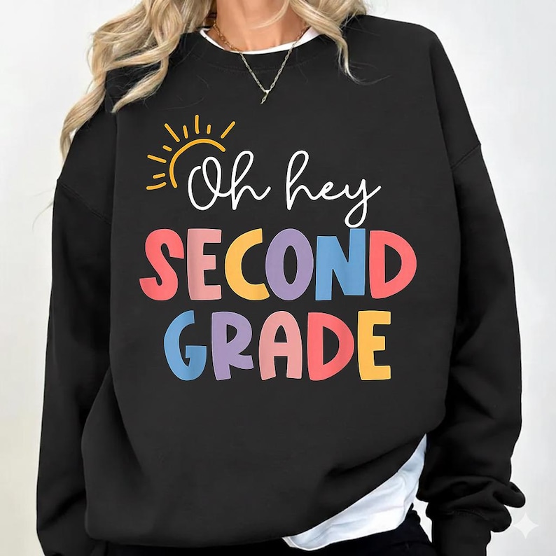 May include: Black sweatshirt with the text "Oh hey SECOND GRADE" in colorful letters. A yellow sun graphic is above the words "Oh hey". The sweatshirt has a white collar and is a casual style.