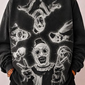 May include: A charcoal grey crewneck sweatshirt featuring a graphic print of five clowns in white face paint. The central clown wears sunflower-shaped glasses and makes a peace sign with both hands, while four other clowns surround it in a circular pattern.