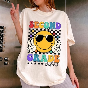 May include: Cream-colored t-shirt with a colorful graphic. The graphic features a yellow smiley face wearing sunglasses and making a peace sign with both hands. The words "SECOND GRADE vibes" are displayed in a retro font.