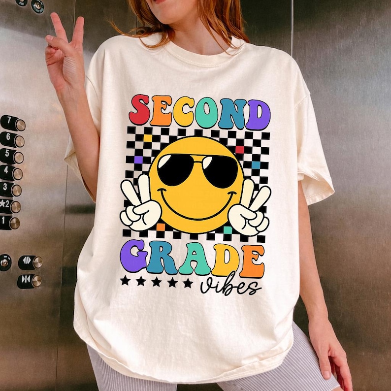 May include: Cream-colored t-shirt with a colorful graphic. The graphic features a yellow smiley face wearing sunglasses and making a peace sign with both hands. The words "SECOND GRADE vibes" are displayed in a retro font.