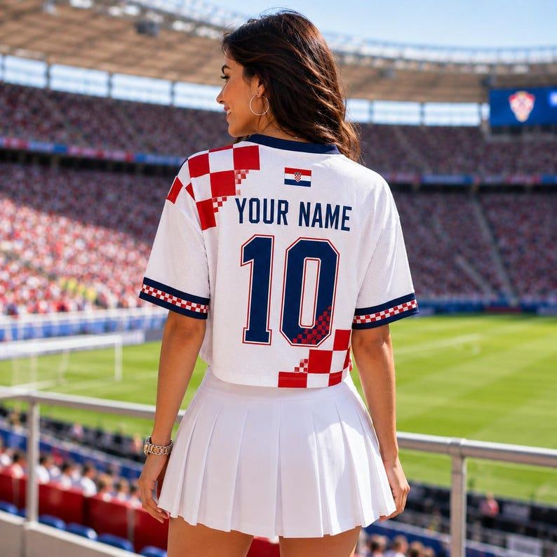 May include: A white cropped jersey featuring a red and white checkered pattern on the shoulder and sleeve, with the text YOUR NAME and the number 10 in dark blue. A small Croatian flag is printed on the back, paired with a white pleated skirt.