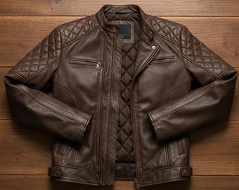 May include: A brown leather jacket with a quilted design on the shoulders and lining. The jacket has a zipper closure, two chest pockets, and a small collar. The jacket is displayed on a wooden surface.