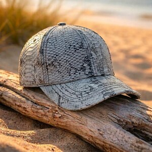 Könnte beinhalten: Eine Baseballkappe mit grau-schwarzem Schlangenledermuster. Die Kappe hat eine strukturierte Krone, einen gebogenen Schirm und sichtbare Nähte an den Paneelen. Die texturierte Oberfläche ahmt das Aussehen von Reptilienschuppen in neutralen Tönen nach.