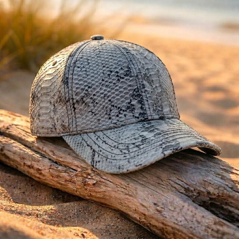 May include: A baseball cap featuring a grey and black snakeskin pattern. The cap has a structured crown, a curved brim, and visible stitching details throughout the panels. The textured surface mimics the appearance of reptile scales in neutral tones.