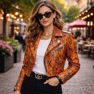 Puede incluir: Chaqueta de cuero naranja y negra con estampado de piel de serpiente, con cinturón negro y hebilla dorada. La chaqueta de estilo motero tiene cremalleras y un corte corto. La persona lleva gafas de sol negras y joyas doradas.