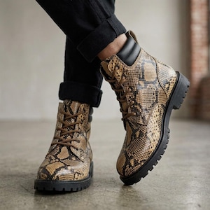Könnte beinhalten: Knöchelhohe Schnürstiefel mit einem beigefarbenen und braunen Schlangenledermuster. Die Stiefel haben einen schwarzen gepolsterten Schaftrand, dunkelbraune Schnürsenkel und eine dicke schwarze Profilsohle. Das Design umfasst Metallösen und eine feste Form.