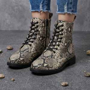 May include: A pair of beige and black snake-print combat boots with black laces and zippers. The boots have a flat sole and are worn with blue jeans.