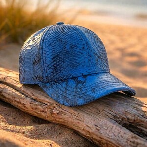 May include: A baseball cap featuring a vibrant blue snakeskin-textured pattern. The cap has a structured six-panel crown, a curved brim, and a consistent scale-like finish across the entire surface, showcasing a detailed reptilian aesthetic.