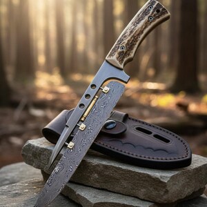 May include: A hunting knife with a patterned blade and a handle made of antler material. The knife is displayed on a stone surface with a brown leather sheath. The blade has a unique design with brass accents.