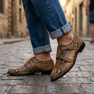 May include: A pair of brown and tan snakeskin-patterned double monk strap shoes with gold-tone buckles, worn with cuffed blue denim jeans. The shoes feature a pointed toe, a low heel, and a textured reptilian print across the entire upper surface.