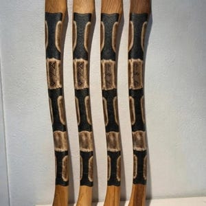 Könnte beinhalten: Vier lange, geschwungene Axtstiele aus Holz, vertikal angeordnet. Jeder Stiel hat eine natürliche hellbraune Holzmaserung mit abwechselnden Abschnitten aus dunklen, strukturierten schwarzen Schnitzereien und glattem Holz. Jeder Stiel zeigt einzigartige Runensymbole.