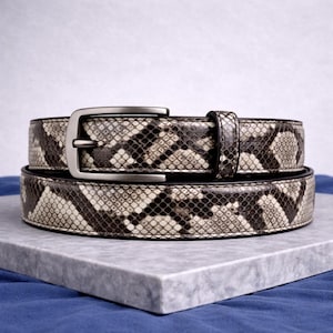 May include: A snakeskin-patterned belt with a silver-colored buckle. The belt is made of a light beige, brown, and black material. The belt is displayed on a gray marble surface against a blue background.