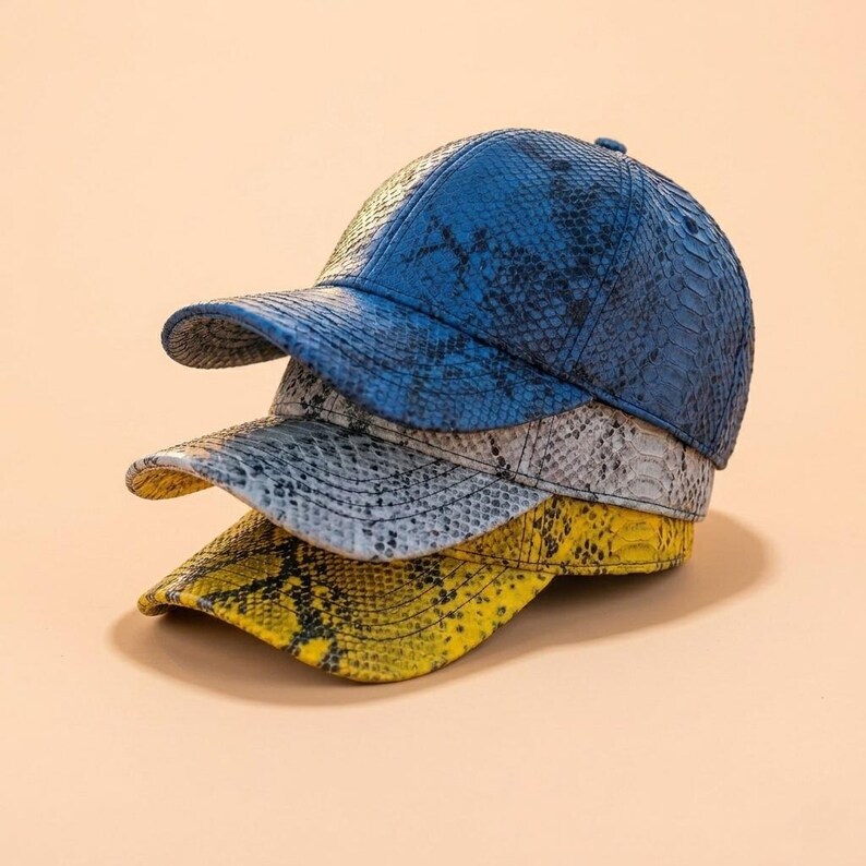 May include: A stack of three baseball caps featuring a textured snakeskin pattern. The top cap is blue, the middle cap is light gray, and the bottom cap is yellow. Each cap has a curved brim and a structured crown with a button on the top.