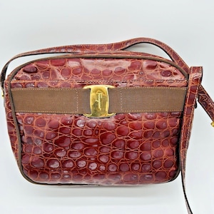 May include: A burgundy-colored crossbody bag with a textured, crocodile-like pattern. The bag features a brown ribbon across the front, a gold-colored buckle, and a matching shoulder strap. The bag is a vintage accessory.