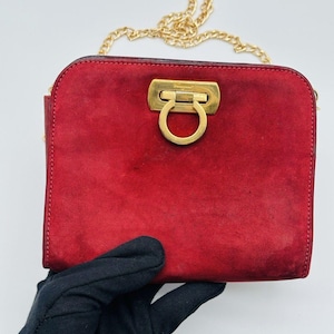 May include: A red suede handbag with a gold-tone chain strap. The bag features a gold clasp with a ring detail. The bag is held by a gloved hand against a white background. The bag is approximately 8 inches wide.