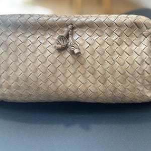 May include: A taupe-colored clutch bag featuring an intricate woven leather pattern across the entire exterior. The rectangular-shaped bag includes a top closure secured with two small, matching leather tassels hanging from the center front.