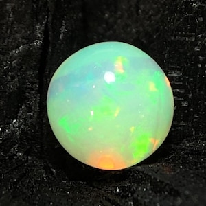 Könnte beinhalten: Ein runder, im Cabochon-Schliff gehaltener Opal-Edelstein mit einer durchscheinenden weißen Basis und lebhaften Blitzen in Neongrün, Gelb und Orange auf der gesamten Oberfläche, vor einem dunklen, texturierten, kohleartigen Hintergrund.