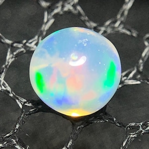 Könnte beinhalten: Ein runder, durchscheinender Opal-Edelstein mit einer glatten, polierten Oberfläche. Der Stein zeigt ein Farbspiel mit internen Blitzen in leuchtendem Grün, zartem Rosa und Blautönen auf einer milchigen, halbtransparenten weißen Basis.