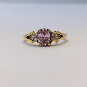 May include: A gold-toned ring featuring a large, oval-cut pink gemstone as the centerpiece. The ring is accented with smaller, clear gemstones on each side of the main stone, set in a decorative design. The ring is a classic jewelry piece.
