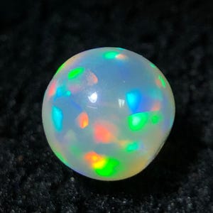 Könnte beinhalten: Ein runder, im Cabochon-Schliff gefertigter weißer Opal-Edelstein mit einer glatten, polierten Oberfläche. Der Stein zeigt ein lebhaftes Farbspiel mit deutlichen Blitzen in Neongrün, leuchtendem Orange und Elektroblau auf seinem Körper.