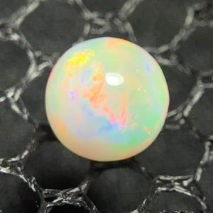 Könnte beinhalten: Ein runder, im Cabochon-Schliff gehaltener Opal-Edelstein mit milchig weißer Grundfarbe und lebhaften orangefarbenen, gelben, grünen und blauen Farbspielen auf der gesamten Oberfläche. Der Edelstein hat eine glatte, polierte Oberfläche und einen Glanz.