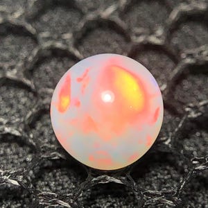 Könnte beinhalten: Ein runder, durchscheinender weißer Opal-Edelstein mit internen Blitzen in leuchtendem Orange, Gelb und zartem Rosa. Die glatte, polierte Oberfläche des Steins reflektiert das Licht und erzeugt einen hellen Glanzpunkt auf der gewölbten Oberseite.