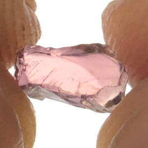May include: A raw, translucent pink gemstone with an irregular, angular shape and faceted surfaces. The stone displays varying shades of light pink and rose, with visible internal inclusions and natural cleavage lines throughout its structure.