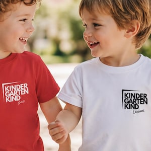 Könnte beinhalten: Zwei Kinder tragen passende T-Shirts. Eines ist rot, das andere weiß. Beide Shirts haben den Text "KINDERGARTEN KIND" in einem schwarzen Quadrat, darunter ein Kindername. Die Kinder lächeln.