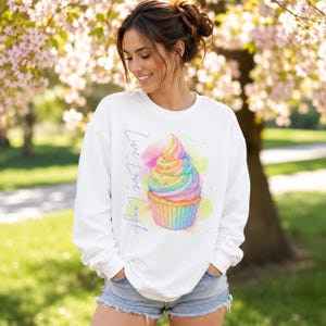 Könnte beinhalten: Weißes Sweatshirt mit einem farbenfrohen Aquarell-Cupcake-Design. Der Cupcake hat Regenbogen-Zuckerguss und eine gelbe Papierform. Die Worte "Live Love Laugh" sind vertikal auf der linken Seite des Sweatshirts aufgedruckt.