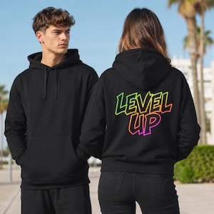 Könnte beinhalten: Zwei schwarze Kapuzenpullover. Einer wird von einer Person getragen, die nach vorne schaut. Der andere Kapuzenpullover hat die Worte "LEVEL UP" in Neon-Grün, Gelb, Orange und Pink auf dem Rücken. Die Kapuzenpullover haben eine Kapuze und eine Vordertasche.