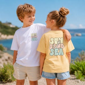 Op de afbeelding: Twee kinderen dragen witte en gele t-shirts met de tekst "SUN, SAND, SEA" in pastelkleuren. Het witte shirt is links en het gele shirt rechts. De kinderen lachen en kijken elkaar aan. De achtergrond is een blauwe oceaan.