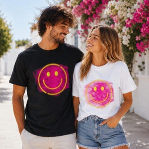 Könnte beinhalten: Ein schwarzes und ein weißes T-Shirt, jeweils mit einem neongelben Smiley-Gesicht-Design auf einem rosa Aquarellhintergrund. Die kurzärmeligen T-Shirts werden von einem Paar getragen, das sich im Freien aufhält.