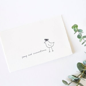 May include: A white card with black ink. The card has a simple drawing of a bird wearing a graduation cap. The text "pomp and circumstance" is written on the card.