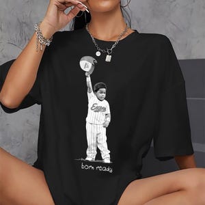 Peut inclure: T-shirt noir avec une illustration en noir et blanc d'un jeune joueur de baseball levant une casquette. Le joueur porte un uniforme à rayures avec le mot "Expos". Le texte "born ready" est imprimé en dessous, créant un design motivant.