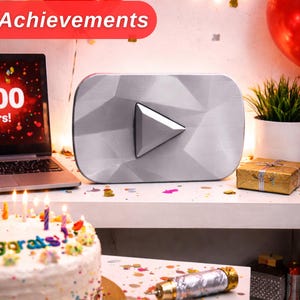 May include: A silver YouTube creator award with a geometric surface, a laptop displaying a 100,000 subscribers milestone, and a white frosted cake with colourful sprinkles and the text Congrats. Red and gold balloons and confetti are present.