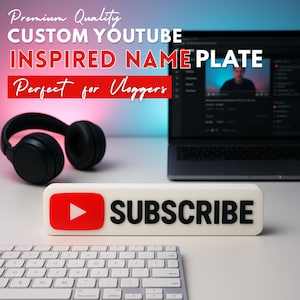 May include: A white rectangular "SUBSCRIBE" button with a red play button icon, a keyboard, and black headphones. The text "Premium Quality CUSTOM YOUTUBE INSPIRED NAME PLATE" is displayed above the button.
