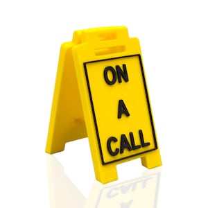 Puede incluir: Un letrero amarillo brillante de dos caras con las palabras "ON A CALL" en negro, encerradas en un borde negro. El letrero es independiente, con un diseño articulado para una fácil configuración y visualización. El letrero está sobre un fondo blanco.