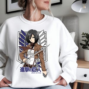 May include: White sweatshirt featuring a graphic of a female anime character with short black hair, wearing a brown jacket and holding a sword. The design includes the text "Attack on Titan" in English and Japanese, with a logo in the background.