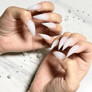 Puede incluir: Manos que muestran uñas acrílicas largas, puntiagudas y blancas. Las uñas son de color blanco opaco sólido y tienen forma de estilete afilado. El fondo es un patrón de mármol claro, con un hilo de perlas.