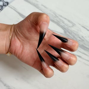 Op de afbeelding: Een hand met lange, puntige acrylnagels. De nagels hebben een gradiëntontwerp, dat overloopt van een natuurlijke basis naar zwarte punten. Een gouden armband met een bedel is zichtbaar op de pols.