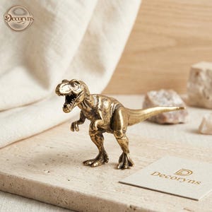 May include: A small, metallic, gold-toned Tyrannosaurus Rex figurine with a textured finish, depicted in a standing position with its mouth open, showing sharp teeth. The dinosaur figure features detailed scales and muscular legs.