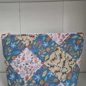 May include: A zipped pouch featuring a patchwork design of diamond-shaped fabric panels. The fabric patterns include blue backgrounds with colourful autumn leaves, white backgrounds with fall foliage, and beige backgrounds with green ribbon bows.