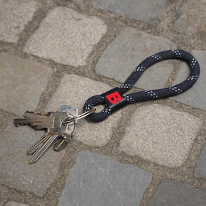 Könnte beinhalten: Ein Schlüsselanhänger mit einer marineblauen und weißen Seilschlaufe und einem roten "B"-Logo. Der Schlüsselanhänger ist an einem Satz Schlüssel befestigt. Der Schlüsselanhänger liegt auf einer Pflastersteinoberfläche.