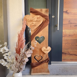 Könnte beinhalten: Ein rustikales Willkommensschild aus Holz. Das Schild hat herzförmige Ausschnitte und Holzherzen mit Text. Ein braunes Band und Trockenblumen ergänzen die Dekoration. Das Schild steht in der Nähe einer Tür.