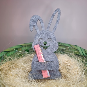 Könnte beinhalten: Graue Filz-Hasen-Dekoration mit einem rosa und schillernden Kamm, platziert in einem grünen Weidenkorb mit Stroh. Der Hase hat ein lächelndes Gesicht und lange Ohren, ideal für Ostern oder Frühlingsdekoration.