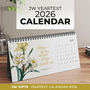 Op de afbeelding: Witte bureaukalender voor 2026 met een bloemenontwerp met gele en witte bloemen. De kalender toont de maand mei en bevat de tekst "Gelukkig zijn zij die zich bewust zijn van hun spirituele behoeften". De kalender staat op een houten oppervlak.