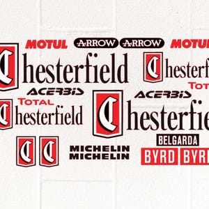 Può includere: Una collezione di loghi e testi, tra cui "MOTUL", "Chesterfield", "ACERBIS", "MICHELIN" e "BYRD". I design presentano schemi di colore rosso, nero e bianco, con vari font e layout. Lo sfondo è una superficie bianca strutturata.