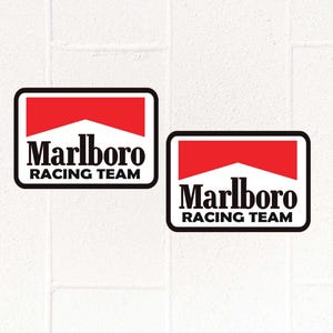 Op de afbeelding: Twee rechthoekige stickers met het Marlboro Racing Team logo. Het logo heeft een rood-wit ontwerp met het woord "Marlboro" boven "Racing Team" in zwarte tekst, alles binnen een zwarte rand.