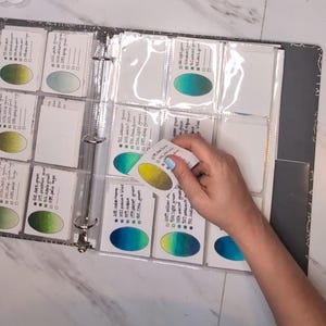 May include: A binder with clear plastic sleeves containing white cards, each featuring a hand-painted oval color gradient and handwritten text labels. One card is being held, displaying a yellow-to-green gradient and ink color names.