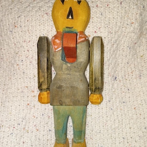 May include: A wooden scarecrow figurine with a pumpkin-shaped head, painted yellow with black triangle eyes and a black stem. The body is gray, with orange and red accents. The arms and legs are painted green and yellow.
